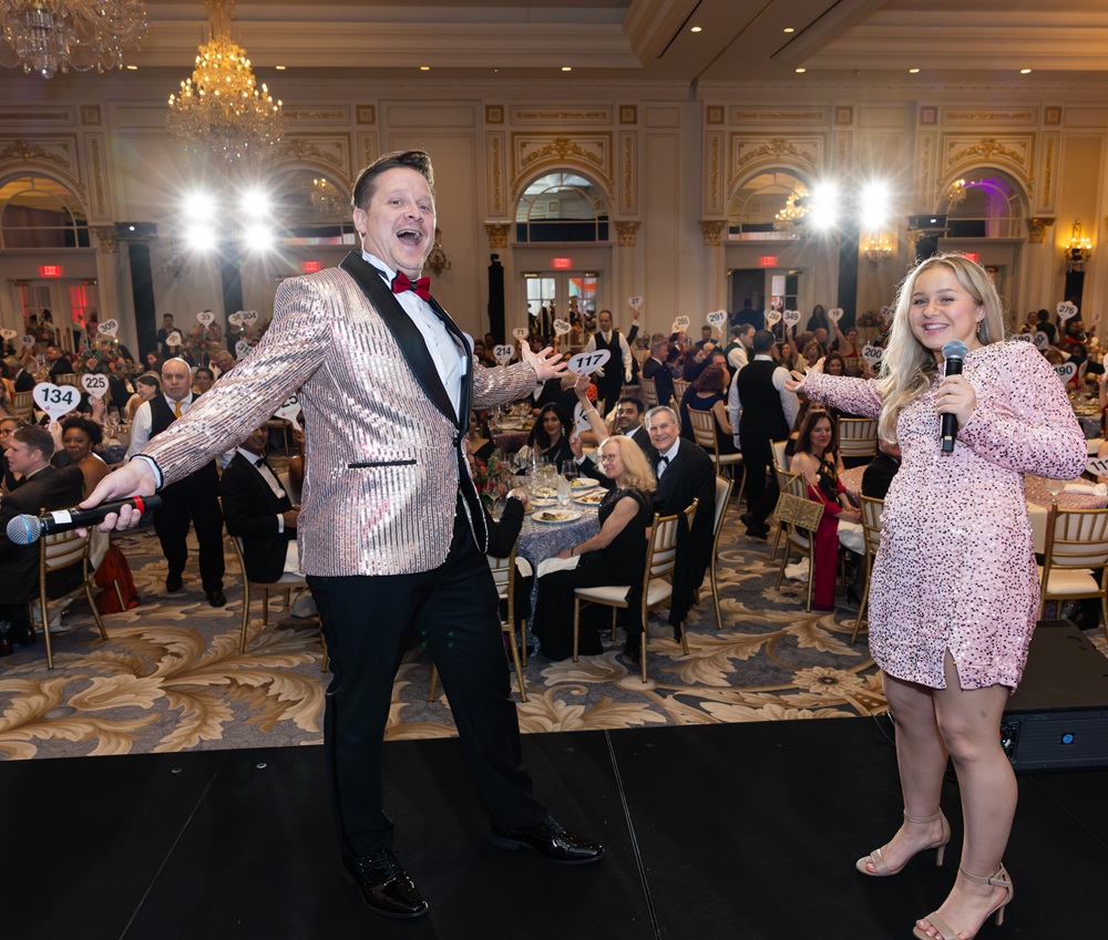 Male and female announcers on stage showing crowd behind them holding up bidder paddles.